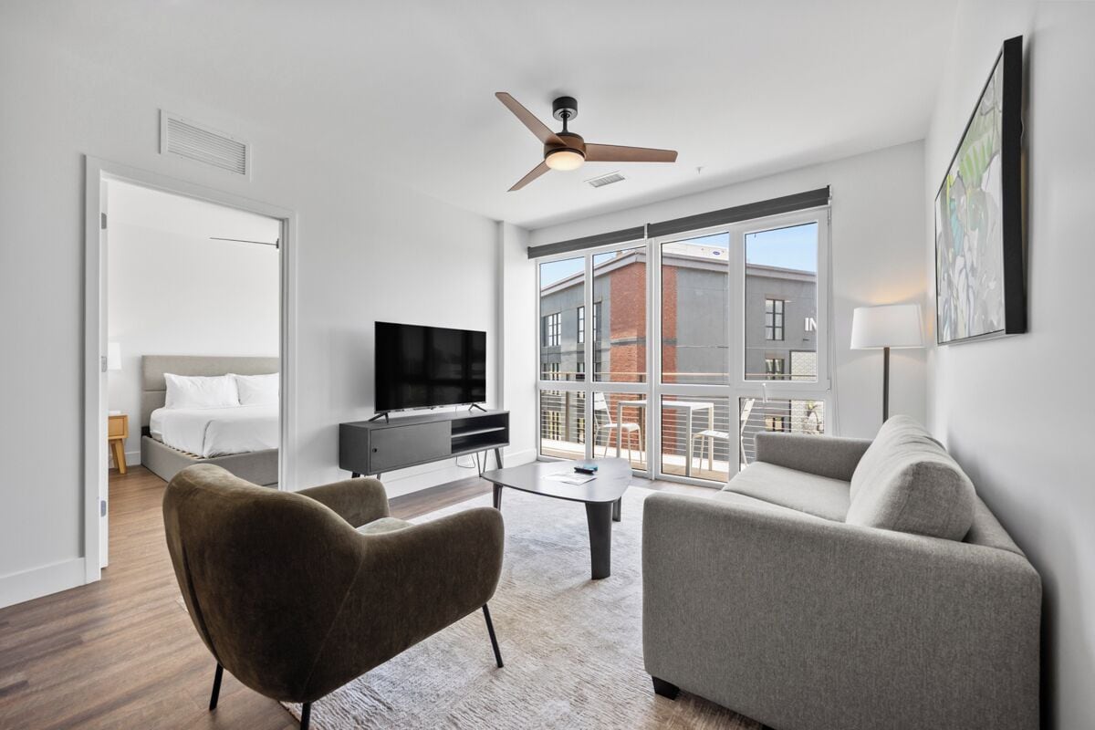 Et moderne hotelværelse med seng, fjernsyn, sofa, sofabord og en balkon med udsigt til bygninger.