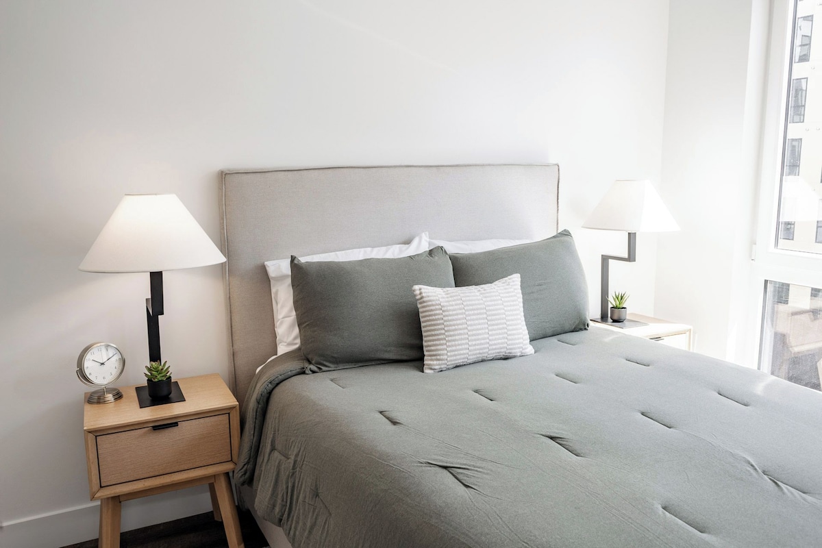 A neatly made bed with grey bedding, two bedside lamps, a small clock, and a plant on a bedside table.