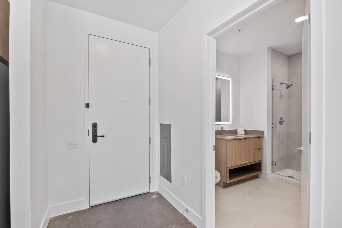 A hallway with a white door, a bathroom with a shower, and a mirror.