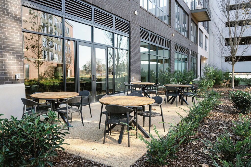 An outdoor seating area with round tables and chairs, surrounded by a garden with various plants and a building with large windows in the background.