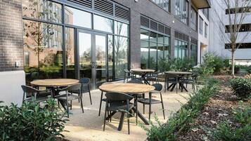 An outdoor seating area with round tables and chairs, surrounded by a garden with various plants and a building with large windows in the background.