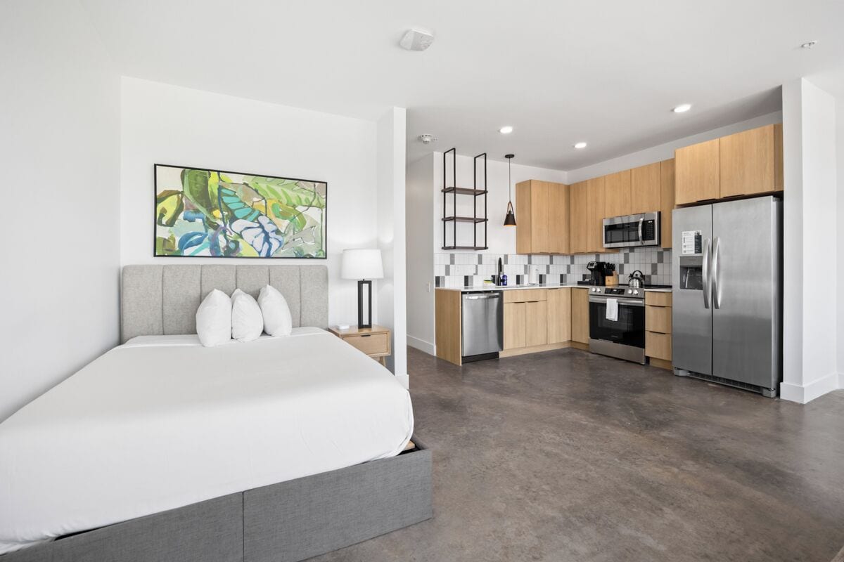 Une chambre moderne avec un grand lit et un tableau accroché au mur. À côté se trouve une cuisine avec des meubles en bois, un réfrigérateur en acier inoxydable et un four à micro-ondes.