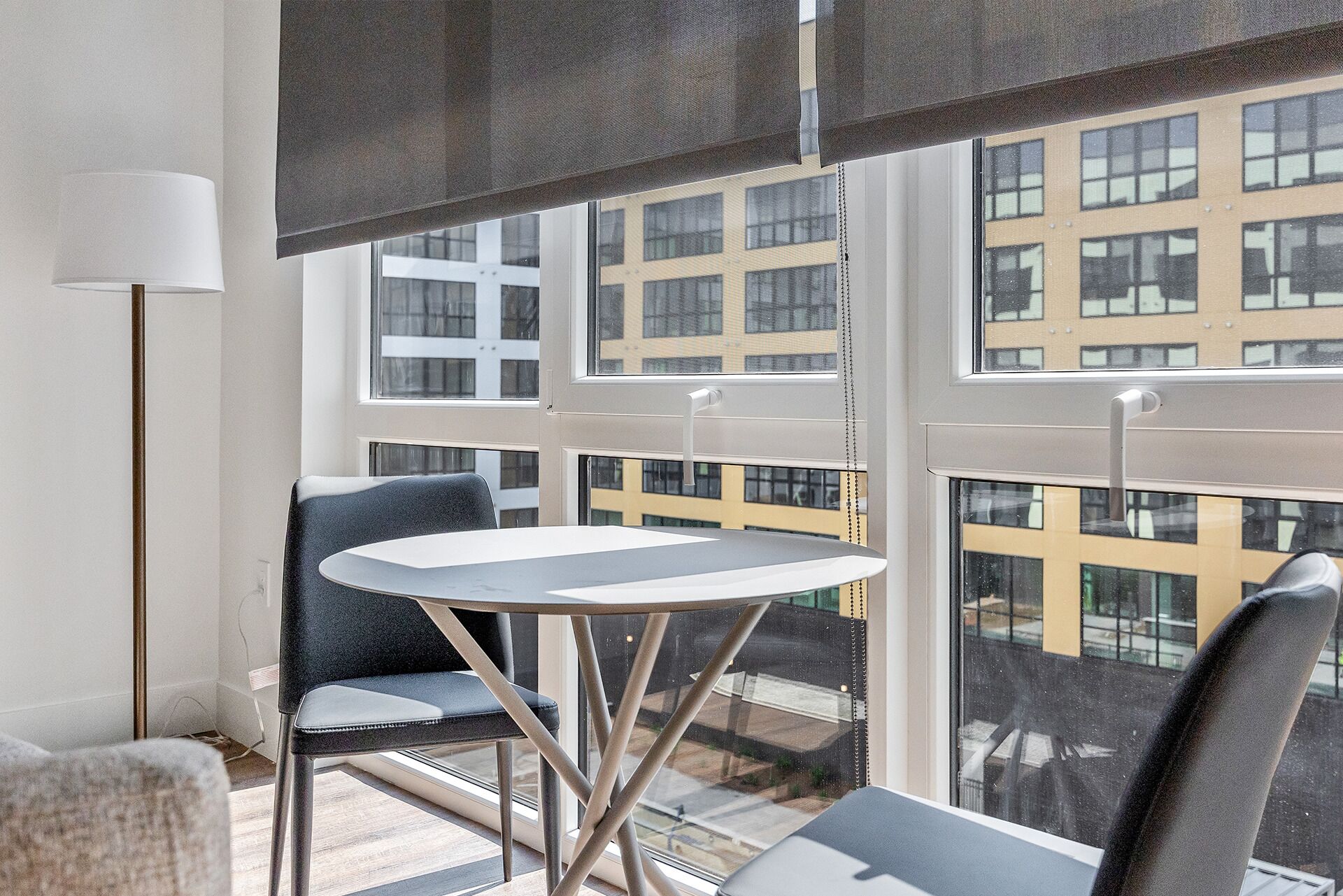 Una habitación moderna con una mesa redonda y dos sillas, ventanas amplias con persianas, y una lámpara de pie.