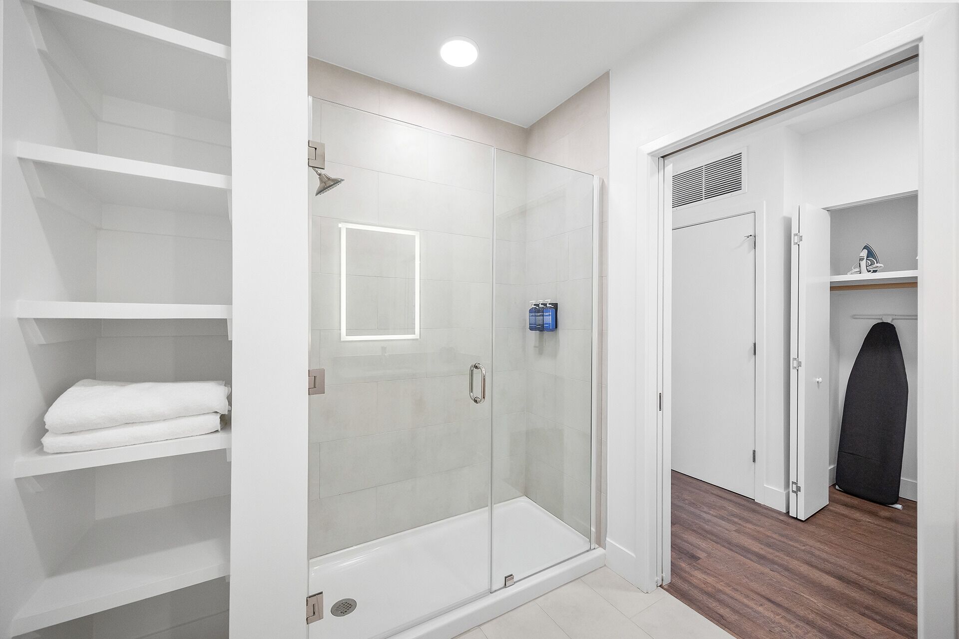 A modern bathroom with a glass shower enclosure, white tiled walls, and a wooden floor.