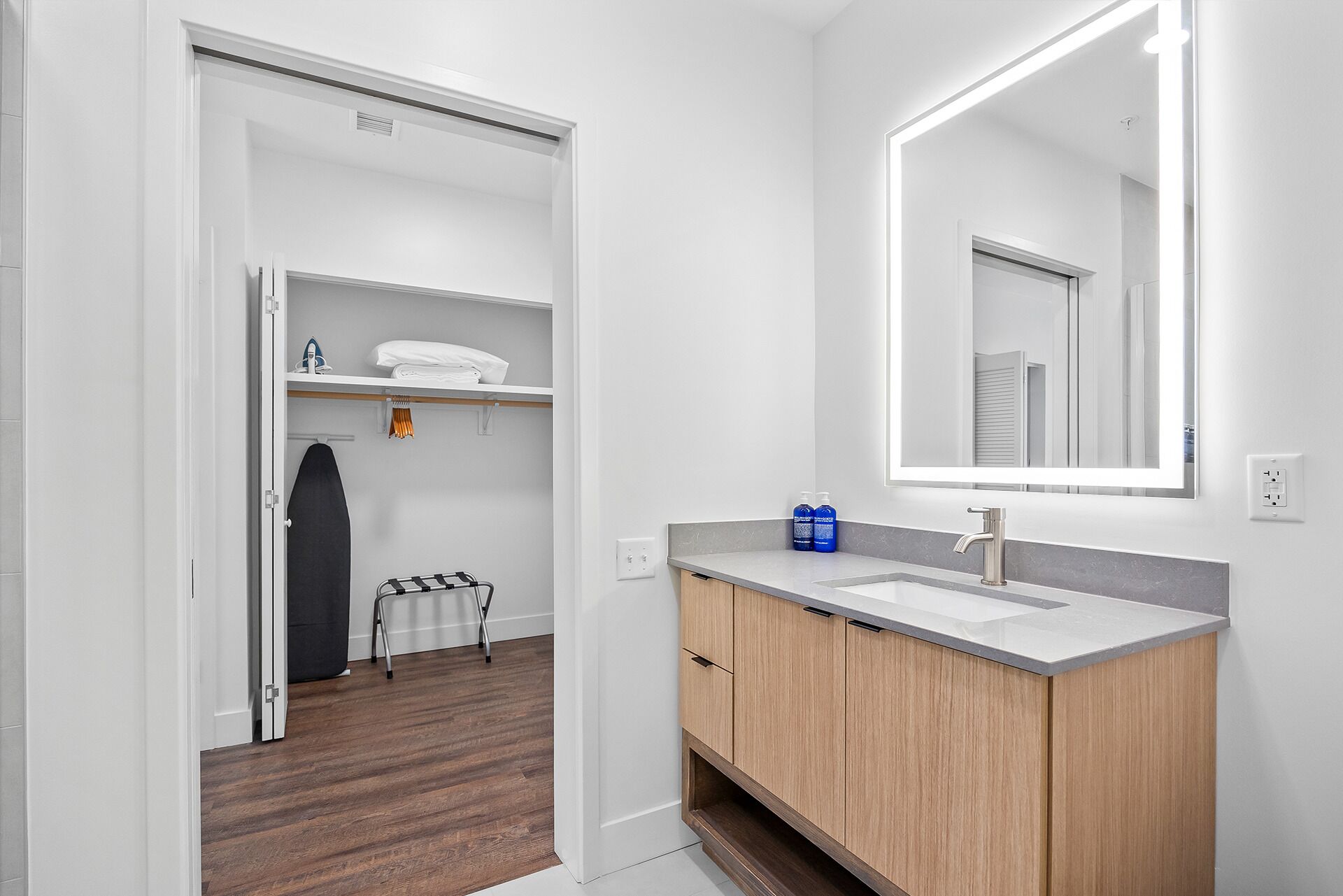 A modern bathroom with a wooden vanity, a large mirror, and a walk-in closet with shelves and a hanging rack.
