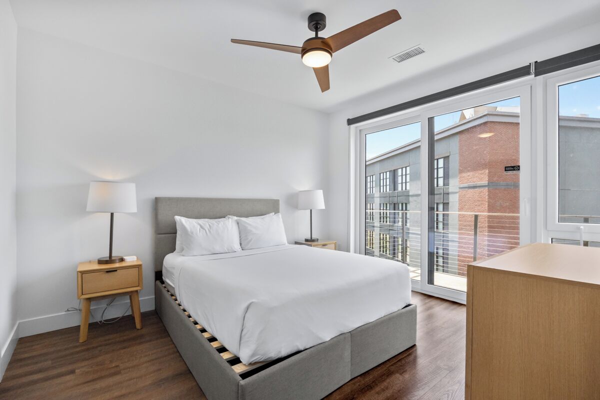 A modern bedroom with a bed, two bedside tables with lamps, a ceiling fan, and a large window with a view of buildings.