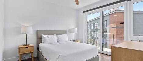 A modern bedroom with a bed, two bedside tables with lamps, a ceiling fan, and a large window with a view of buildings.