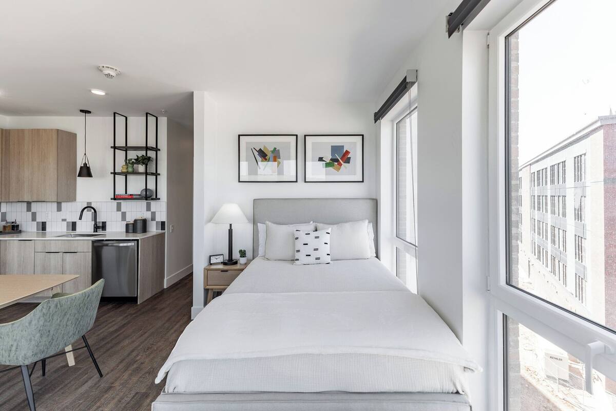 Une chambre d’hôtel moderne équipée d’un lit, d’un bureau et d’une kitchenette. Deux œuvres d’art encadrées ornent le mur, et une grande fenêtre offre une vue sur la ville.