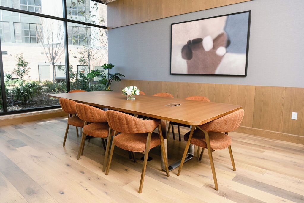 A conference room with a long wooden table, six orange upholstered chairs, and a framed artwork on the wall.