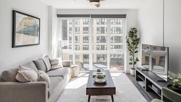 A modern living room with a sofa, coffee table, and TV, large windows with a view of buildings, and a ceiling fan.