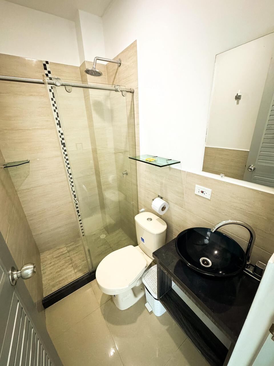 Uma casa de banho moderna com cabine de duche em vidro, sanita branca, lavatório preto e prateleira de vidro.