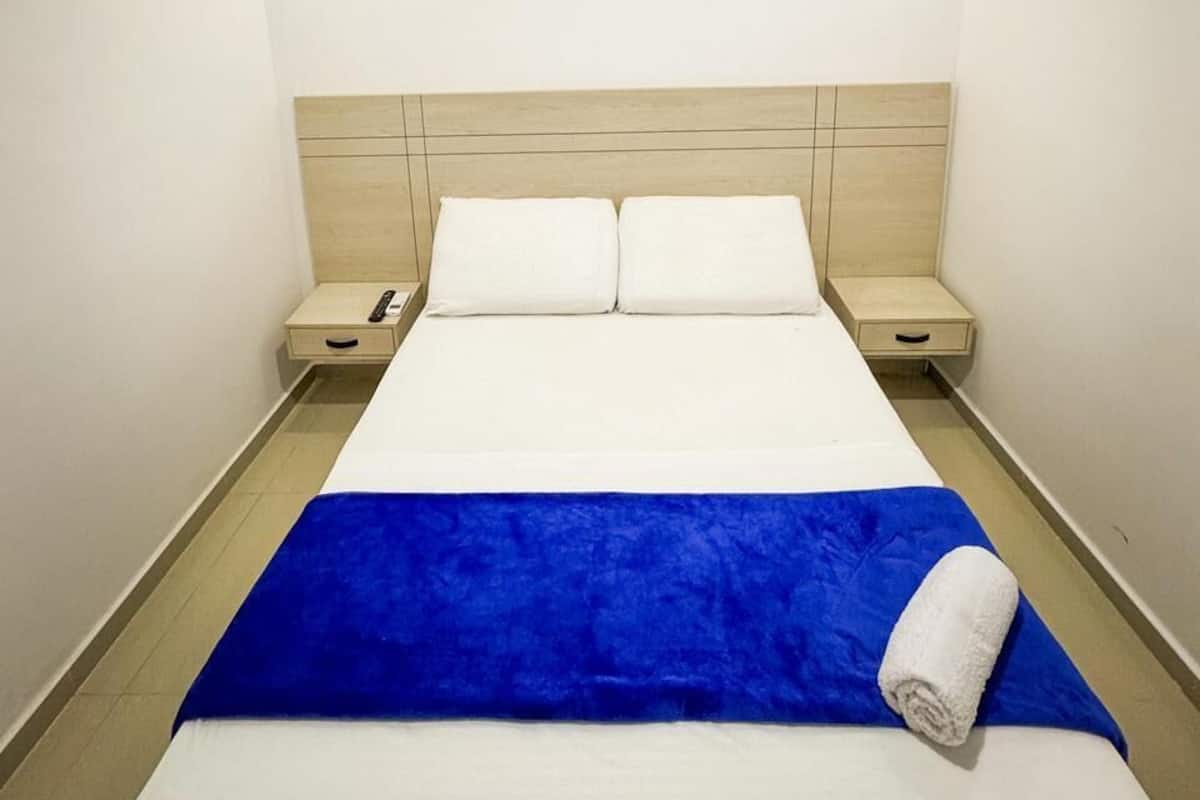 Quarto de hotel com uma cama, um cobertor azul, um travesseiro branco, uma mesinha de cabeceira e um ar-condicionado fixado na parede.