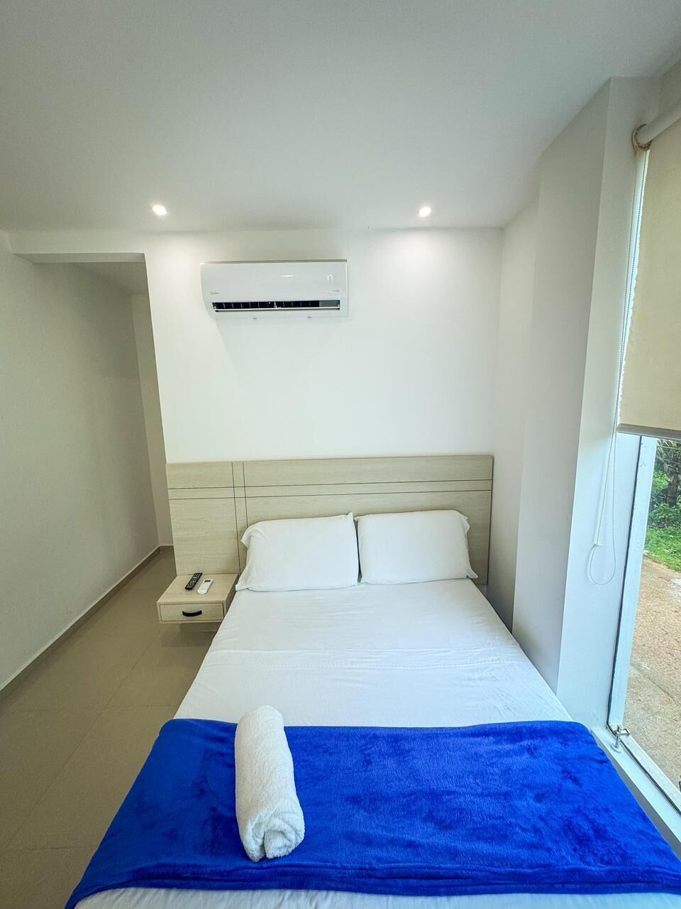 A hotel room with a bed, a nightstand, a remote control, a blue towel, and an air conditioning unit.