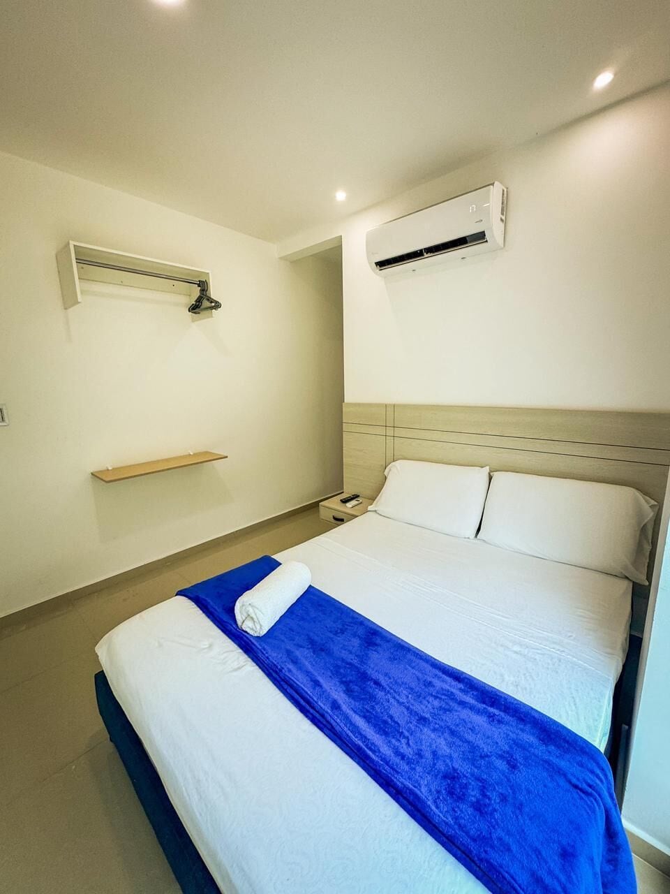 A hotel room with a bed, a nightstand, an air conditioning unit, and a shelf.