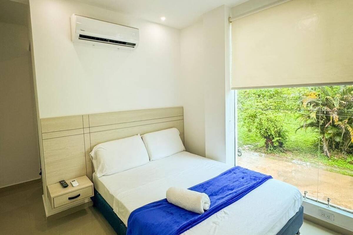 Quarto com cama, mesa de cabeceira, ar condicionado e janela com vista para área verde.