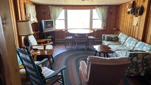 Interior acolhedor de uma cabana, com decoração em madeira, uma mesa de jantar redonda, um sofá e uma televisão.
