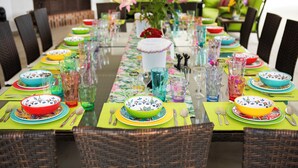 Une table à manger dressée avec des bols, des assiettes et des verres de couleurs vives, entourée de chaises en osier.