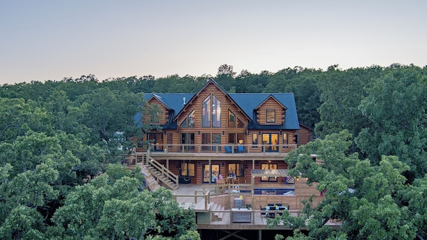 A large wooden cabin with multiple decks and a swimming pool, surrounded by dense forest.