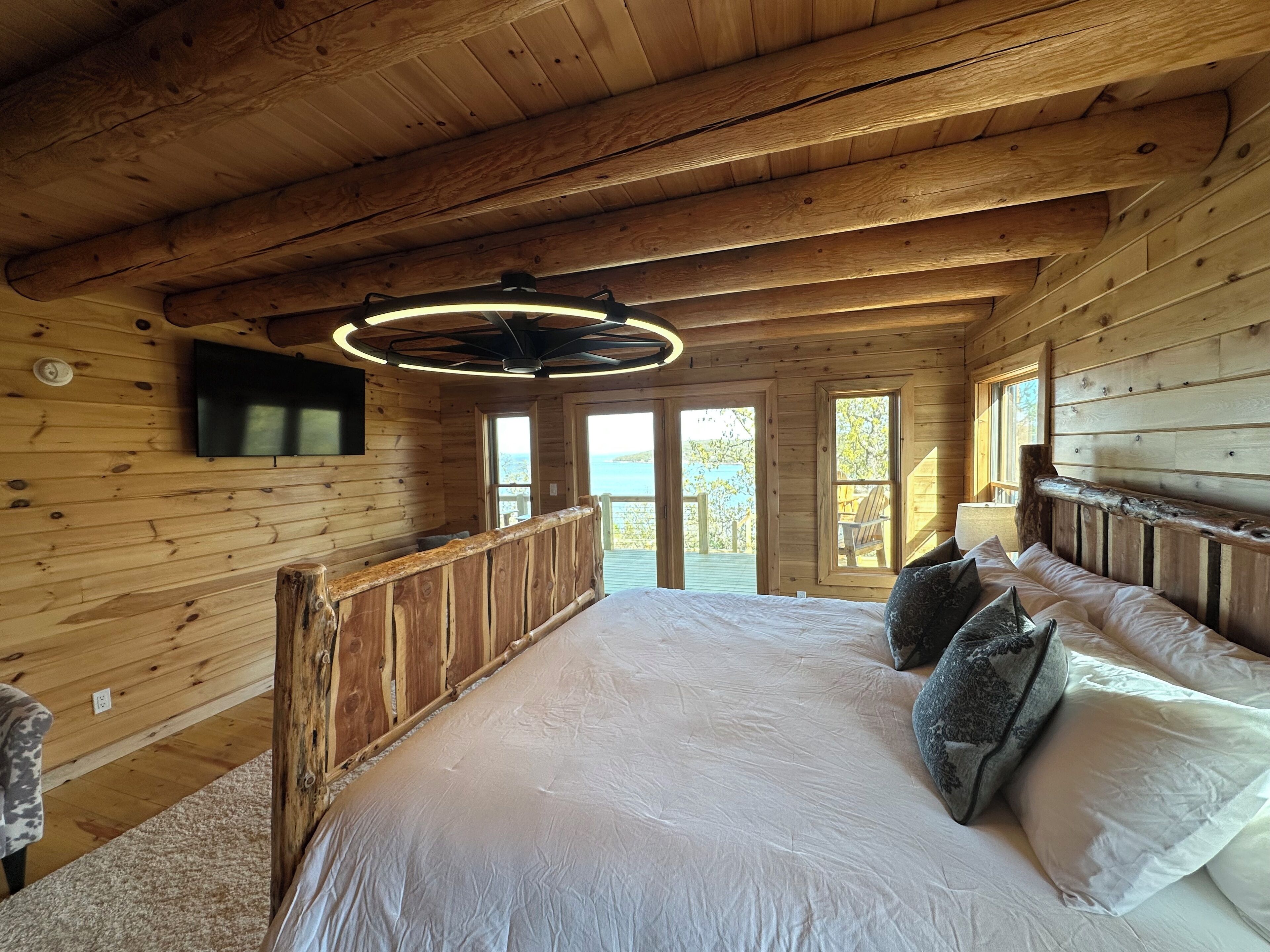 Ein Schlafzimmer einer Holzhütte mit einem großen Bett, einem an der Wand montierten Fernseher und Blick ins Freie durch die Fenster.