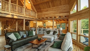 A spacious wooden cabin interior with a living area featuring a sofa, armchairs, and a coffee table. A staircase leads to an upper deck with a railing.