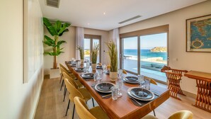 A dining area with a long table set for four, large windows with a view of the sea, and a large plant on the left.