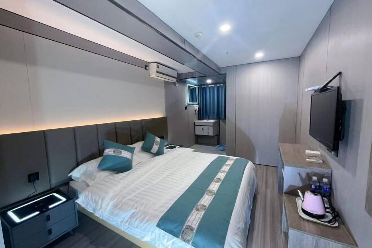 A modern hotel room with a bed, bedside table, TV, and a small kitchenette area.