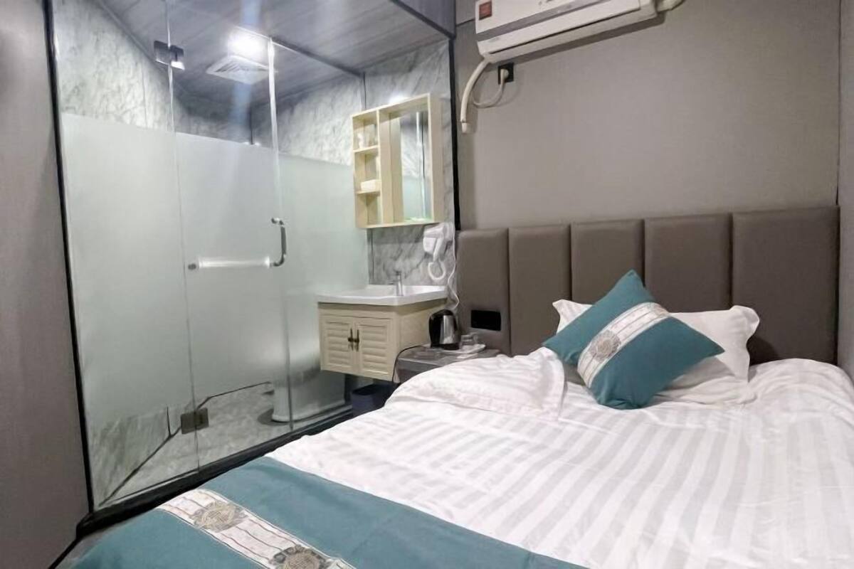 A hotel room with a bed, a glass shower enclosure, a vanity with a mirror, and a wall-mounted air conditioning unit.