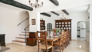 Zona pranzo con tavolo in legno apparecchiato per sei, circondato da sedie. Si vede una scala con ringhiera in legno che conduce a un livello superiore. Un lampadario pende dal soffitto e sullo sfondo è visibile un mobile con piatti.