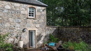 A stone building with a door and window, surrounded by a garden with plants and a wooden bench.