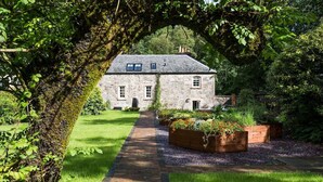 A stone house with a paved pathway leading to the entrance, surrounded by lush greenery and flowering beds.