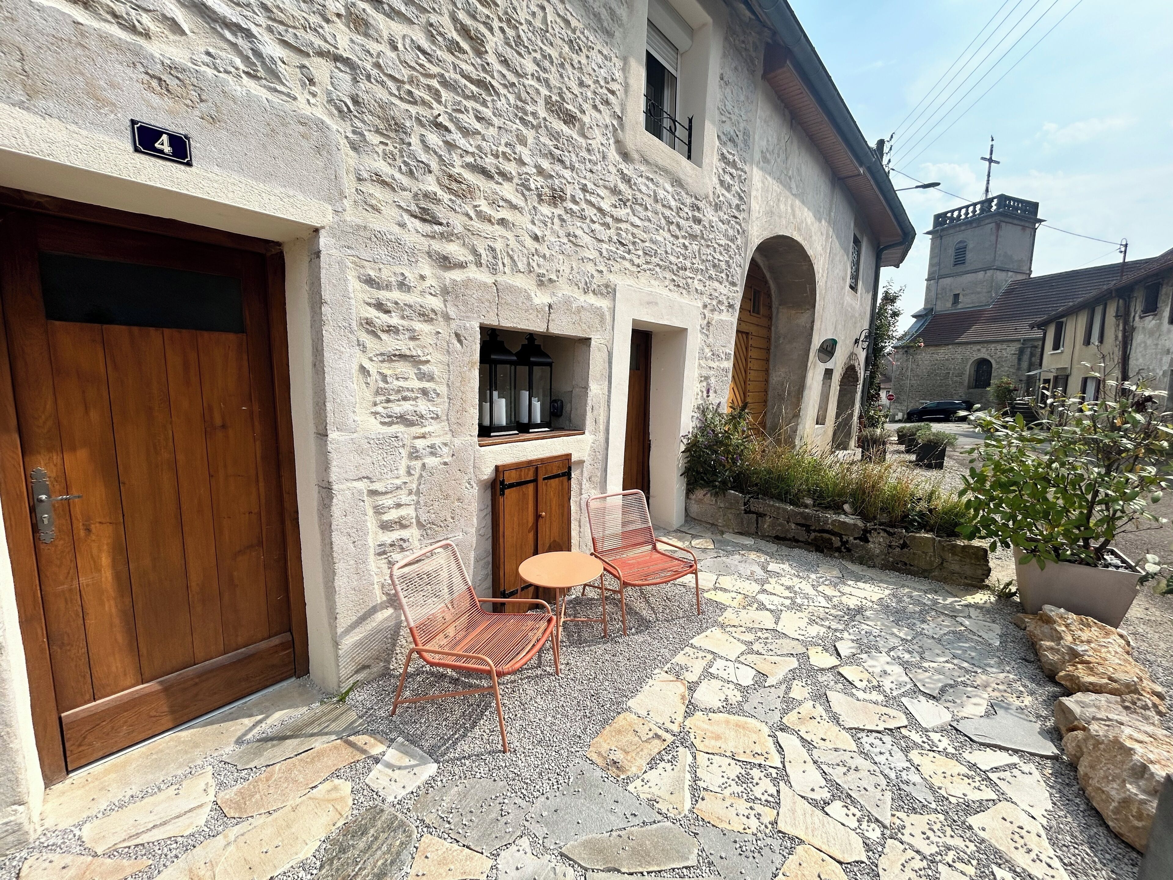 A stone building with a wooden door, a small patio with a table and chairs, and a view of a church in the background.