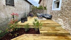 A courtyard with a wooden deck, a stone wall, a grey sofa, and a small table. - Ferme Gabriela | Luxury accommodation in the lake region in the heart of the Jura mountains (Châtillon)