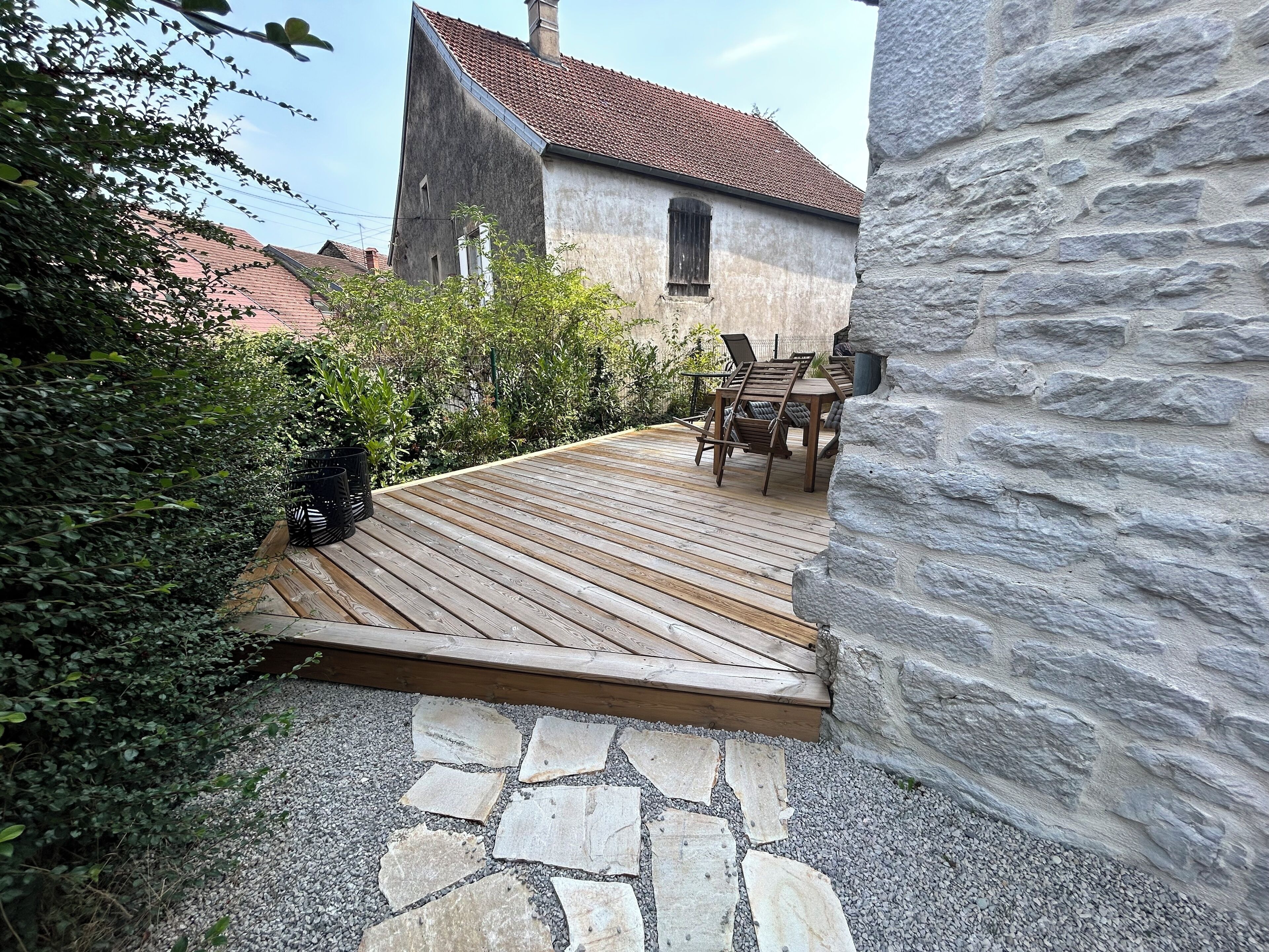 Une terrasse en bois, recouverte de carreaux de pierre, mène à un mur de pierre, le tout étant entouré de verdure et bordé par un bâtiment dont le toit est recouvert de tuiles rouges.