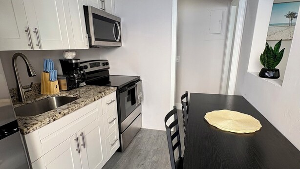 A compact kitchen with white cabinets, a microwave, a stove, and a sink. There's a dining table with chairs and a small potted plant on the wall. - Cozy 1-bed apt in fabulous Pompano Beach across from the beach, AC, WiFi, pets (Pompano Beach)