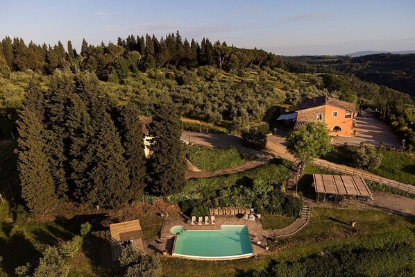 An aerial view of a property with a swimming pool, a stone building, and a forested hillside.