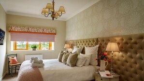 Une chambre avec une tête de lit capitonnée, une grande fenêtre avec des stores, une table de chevet avec une lampe et un fauteuil.