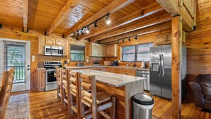 A log cabin-style kitchen with wooden beams, a central island with stools, stainless steel appliances, and a view of the outdoors through large windows. - Hot Tub, Fire Pit, WiFi, EV Charger | Biggie cabin near Red River Gorge, KY! (Stanton)