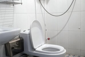 Des toilettes blanches avec un abattant fermé, un bidet et une pomme de douche dans une salle de bain aux murs recouverts de carreaux blancs.