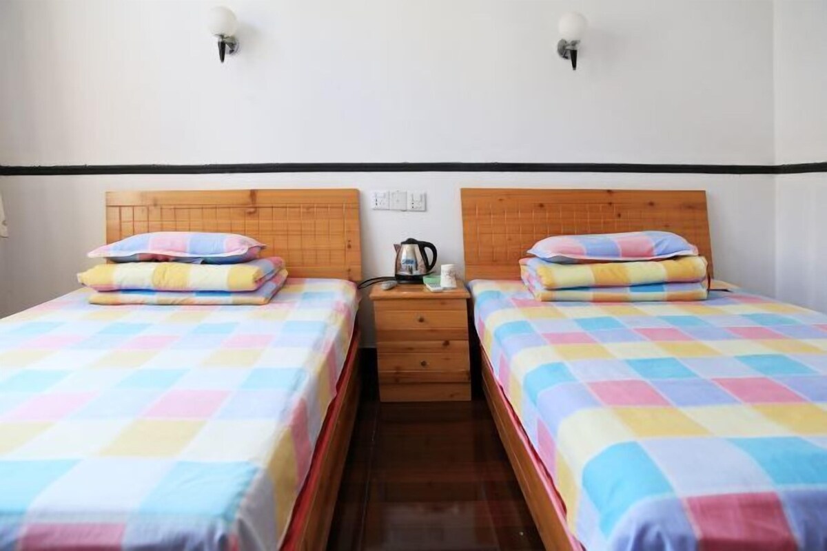 A room with two beds, each with a wooden headboard and a colorful checkered bedspread. There is a nightstand between the beds with a kettle and a cup.