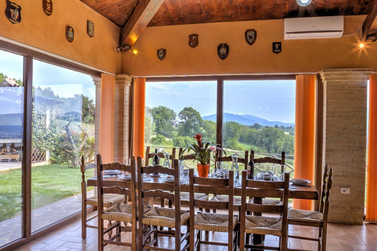 Ein Essbereich mit einem Tisch für vier Personen, große Fenster mit Blick auf eine üppige Landschaft und eine Holzbalkendecke mit dekorativen Wappen.