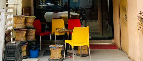 A small outdoor seating area with wicker chairs and tables, a car parked in front, and a building entrance with a glass door.