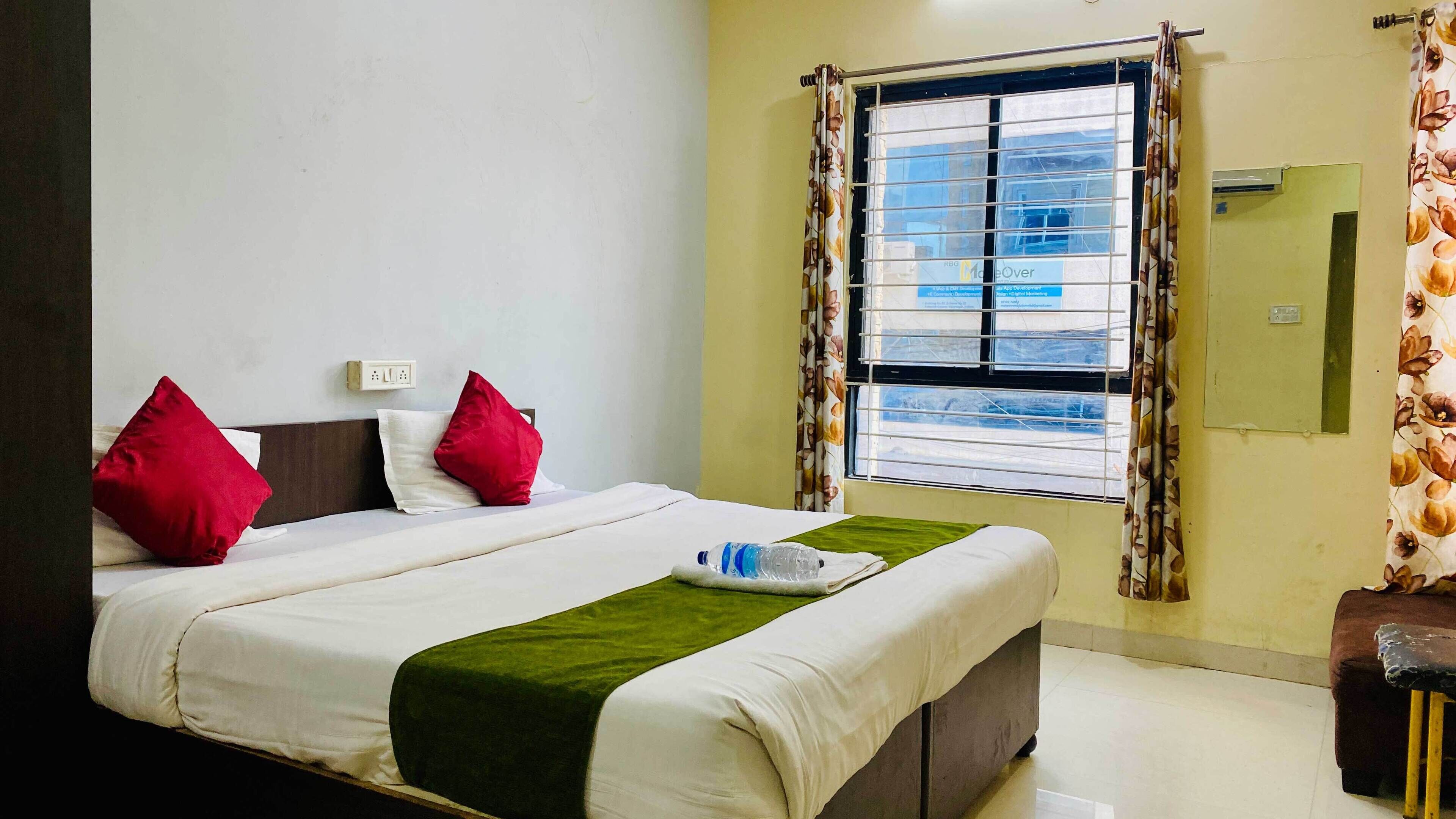 A hotel room with a bed, two pillows, a green throw, a window with blinds, and a small table with a bottle.