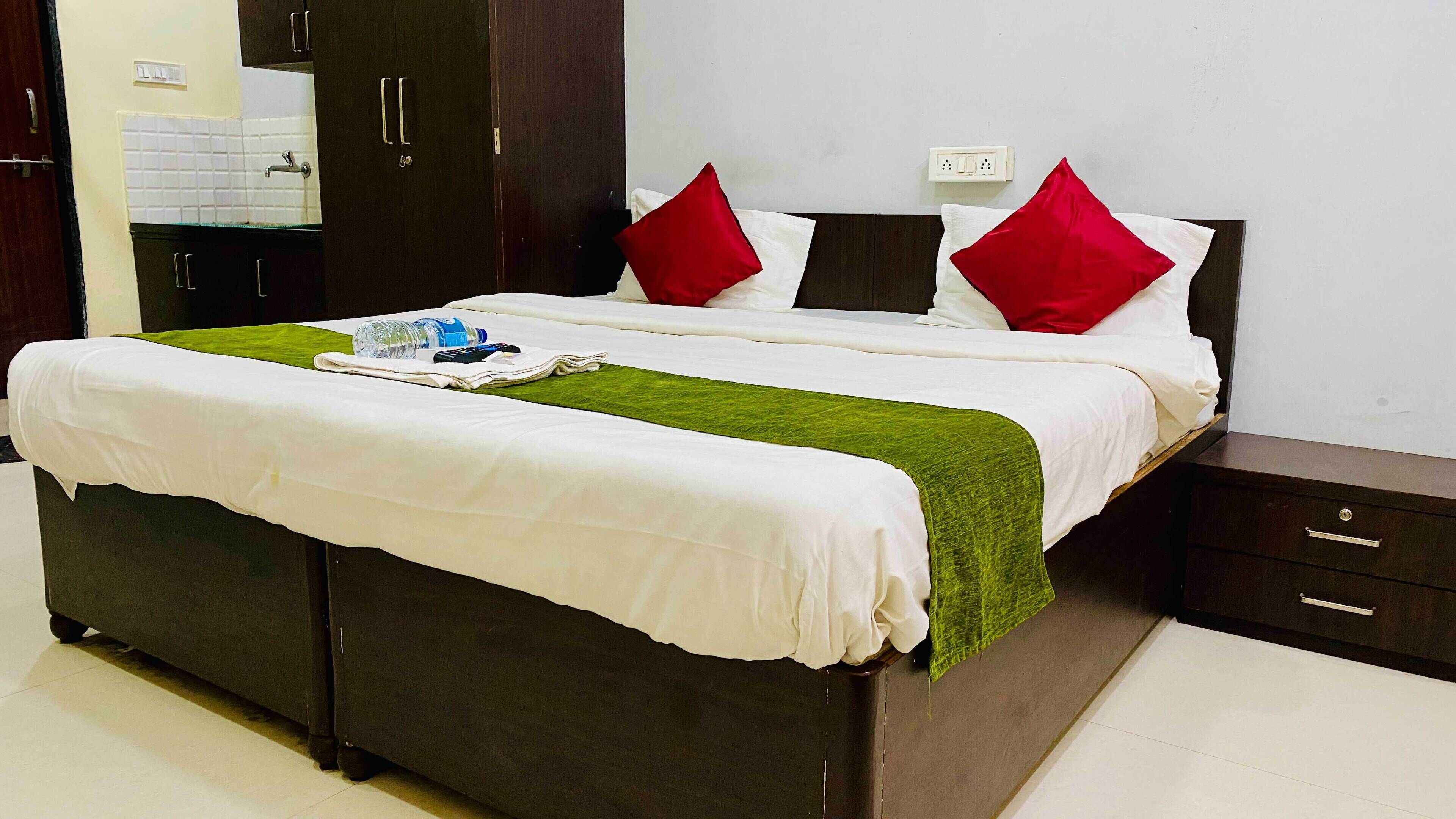 A double bed with white bedding and red pillows, a green throw blanket, and a bedside table with a drawer.