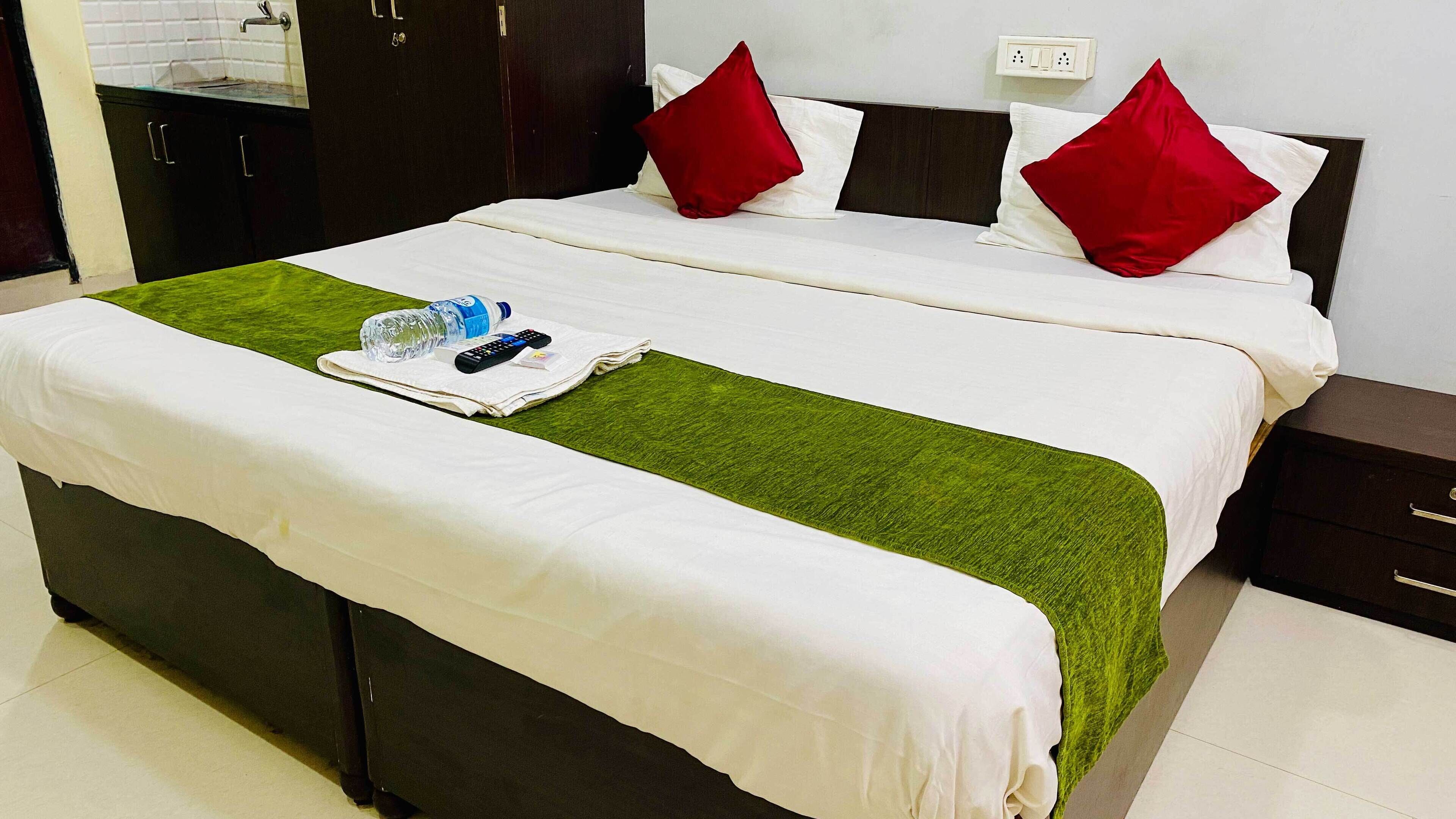 A neatly made bed with white bedding, two red pillows, and a green throw. A remote control and a bottle of water are on the bed.