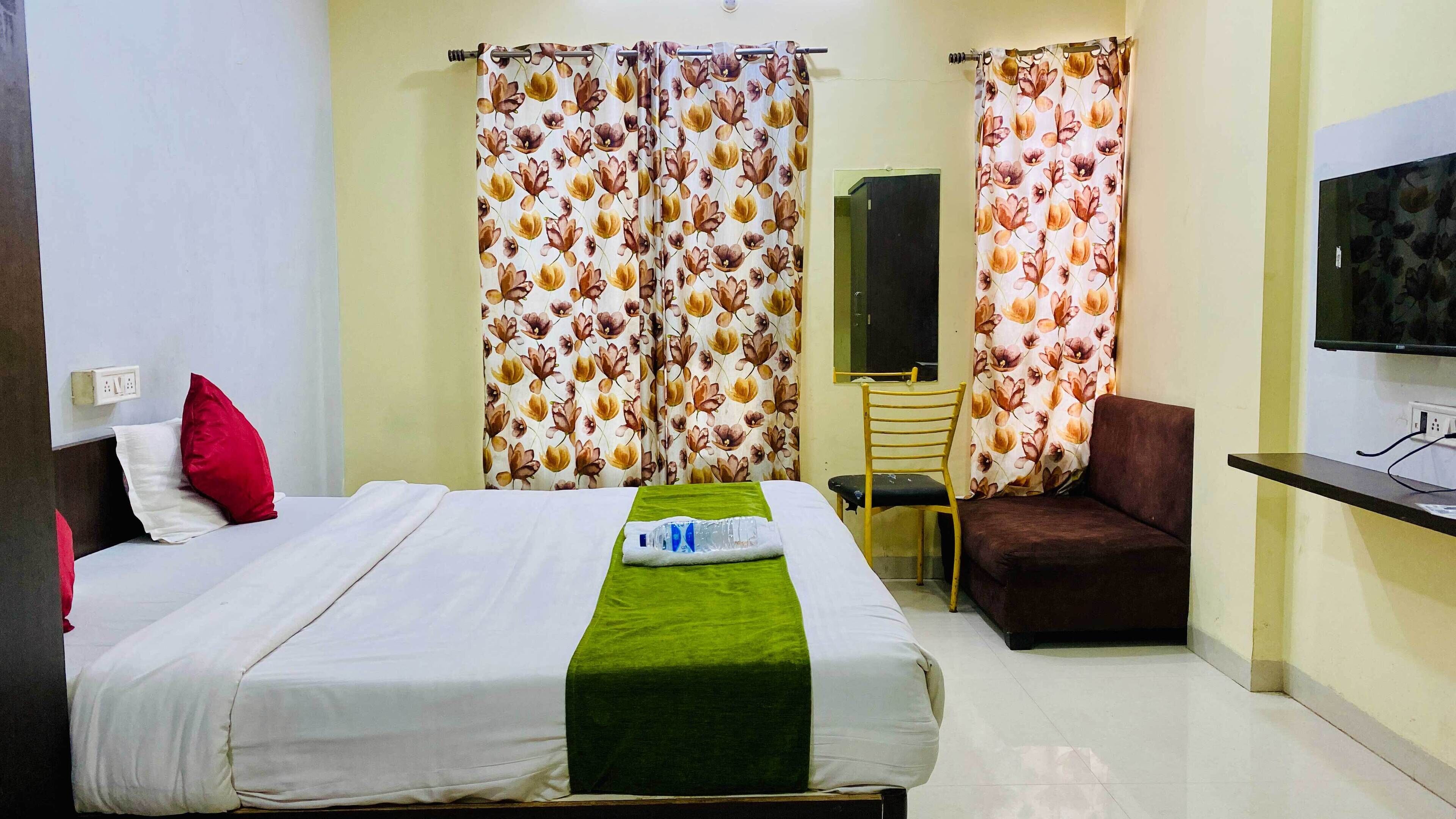 A hotel room with a bed, a green towel, a chair, a sofa, a TV, and floral curtains.