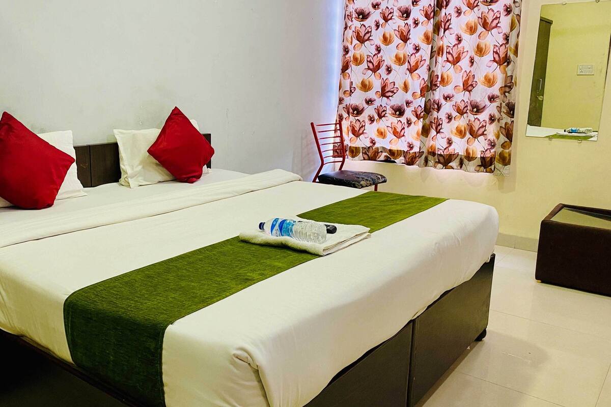 A hotel room with a bed, pillows, a green throw, a water bottle, and a small table.