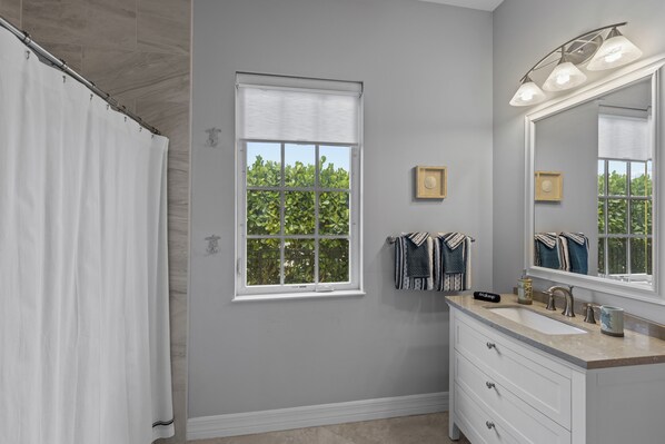 A bathroom with a shower, a sink with a mirror, and a window with a view of greenery. - Sunrise Island Retreat (Fort Pierce)