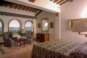 A room with a bed, a desk, two chairs, a dresser, and a large mirror. There are three arched windows offering a view of the landscape.
