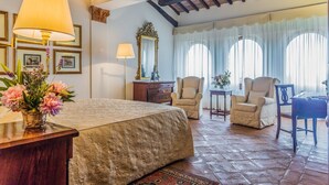 A spacious bedroom with a large bed, two armchairs, a wooden desk, and a mirror.