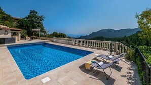 A rectangular swimming pool with clear blue water, surrounded by beige tiles. There are two lounge chairs with towels, a small table, and a pergola with a table underneath. The pool is adjacent to a white building with a balcony, and the area is enclosed by a black metal railing.
